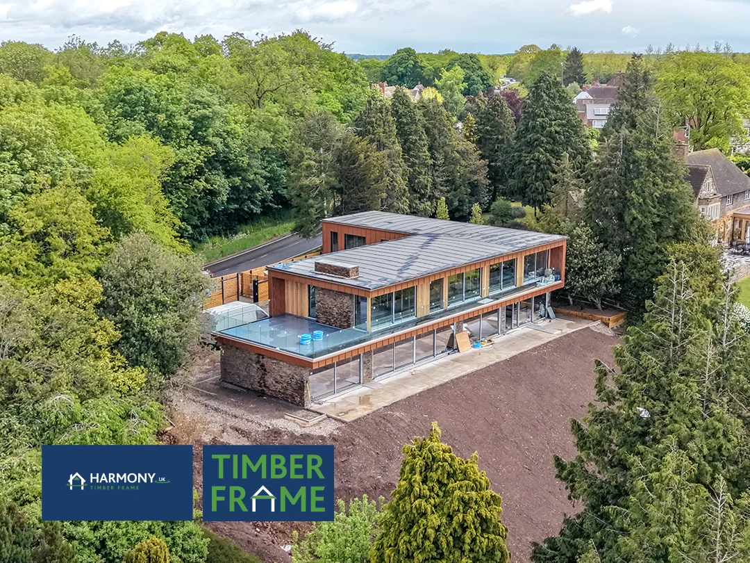 Two-story timber-framed modern house with large glass walls and flat roofs, set among trees with Harmony Timber Frame logo in bottom left corner.