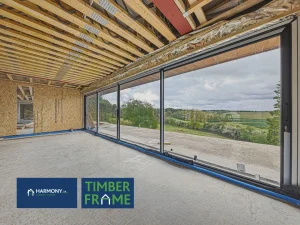 Timber-frame interior with exposed joists and large sliding glass doors overlooking countryside; Harmony Timber Frame logo in bottom left.